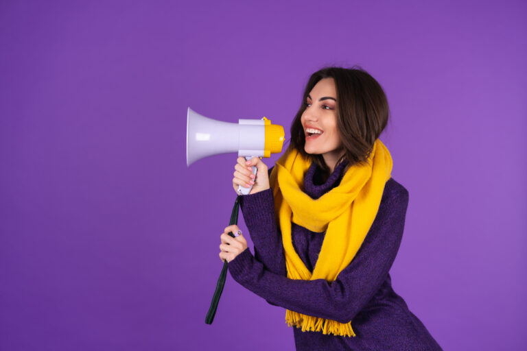 Young woman in a knitted dress and in a yellow scarf on a purple background cheerfully shouts into a megaphone, cheerful, announces a sale